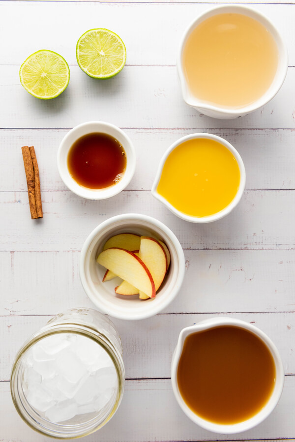 Kitchen table with a container filled with apple cider, another one with orange juice,  another with honey, another one with ginger beer, some limes, a cinnamon stick, apple slices, and  jug with ice cubes