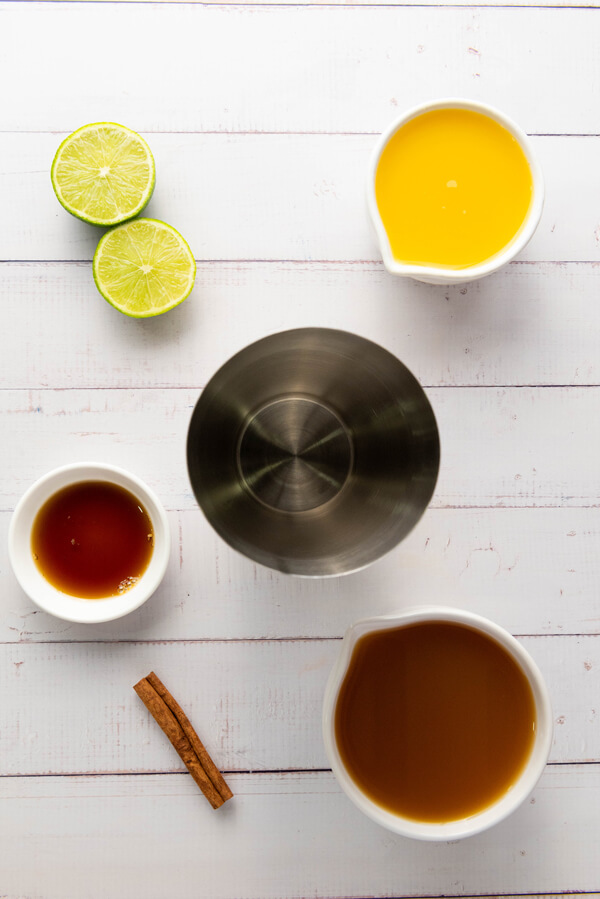 Shaker surrounded by apple cider, orange juice, lime juice, honey, and a cinnamon stick