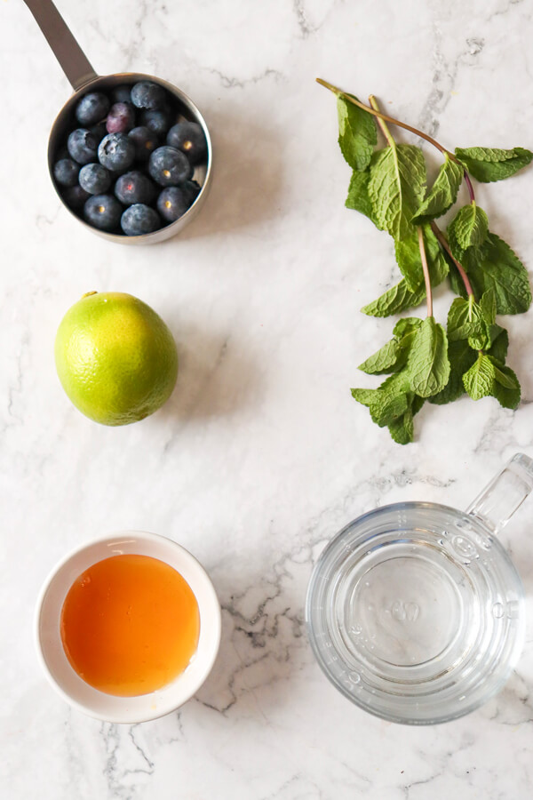Kitchen counter top with a cup of blueberries, mint leaves, a lime, a cup of honey, and a jug of sparkling water