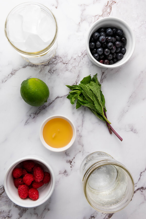 Kitchen counter top with a bowl of blueberries, a bowl with raspberries, a cup of agave syrup, a cup of soda water, a cup of ice cubes, mint leaves, and a lime