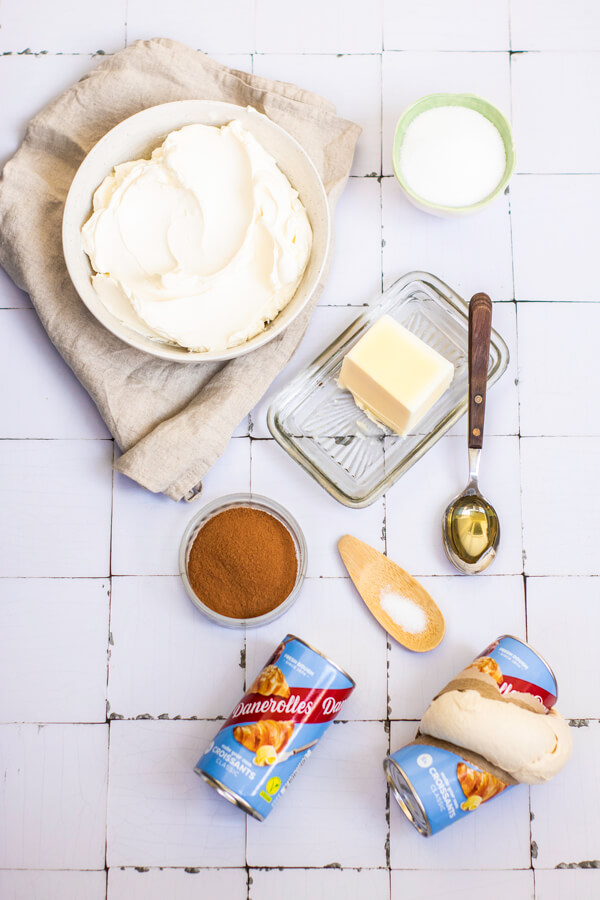 Kitchen counter top with bowls of cream cheese, sugar, salt, and cinnamon, next to a bar of butter, a spoon of vanilla extract, and 2 cans of Crescent Roll Dough