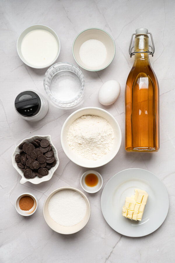 Kitchen counter top with bowls of water, butter, sugar, salt, flour, vanilla extract, cinnamon, heavy cream, and chocolate chips next to one egg and a bottle of oil