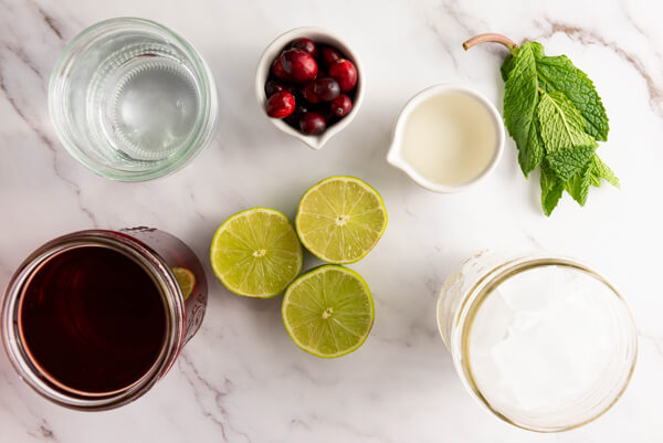 Kitchen counter top with cranberries, cranberry juice, sparkling water, simple syrup, sliced limes, mint leaves, and a bucket of ice