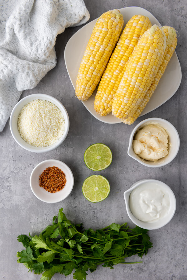 Counter top with bowls of elotes, cream, cotija cheese, mayonnaise, and tajín next to some limes and cilantro