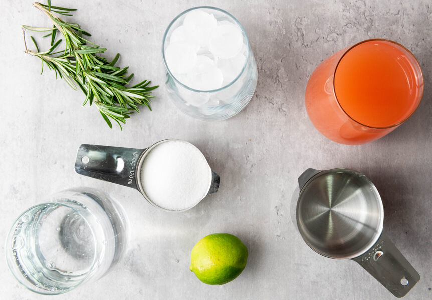 Kitchen counter top with grapefruit juice, sugar, water, rosemary, limes, ice cubes on a glass, and sparkling water