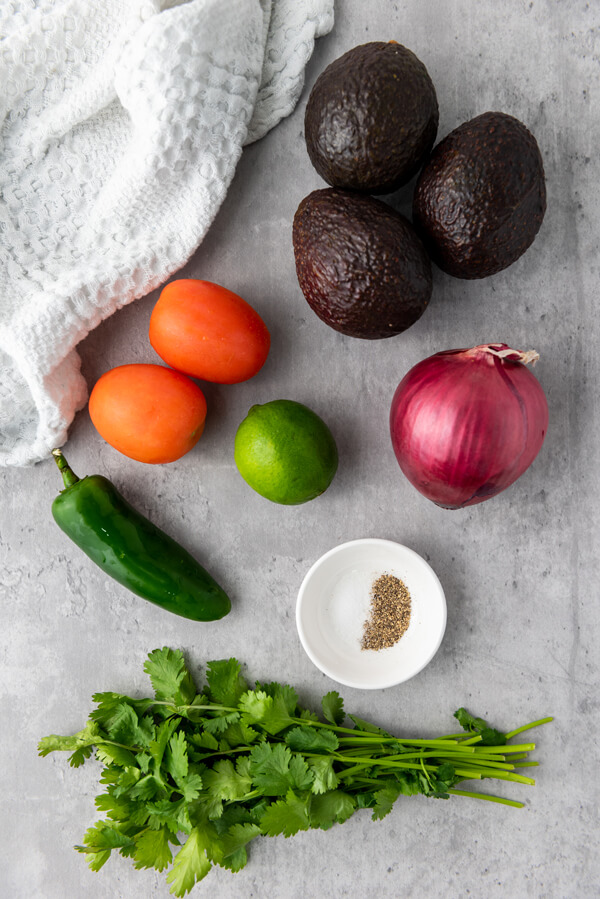 Counter top full of veggies: 3 avocados, 2 tomatoes, 1 onion, 1 jalapeño, 1 lime, and cilantro next to a dish with salt and pepper