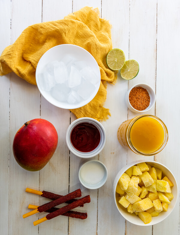 Wooden table with bowls filled with frozen mango cubes, ice cubes, mango nectar, granulated sugar, chamoy sauce, and tajin next to a fresh mango, limes, and tamarind candy sticks