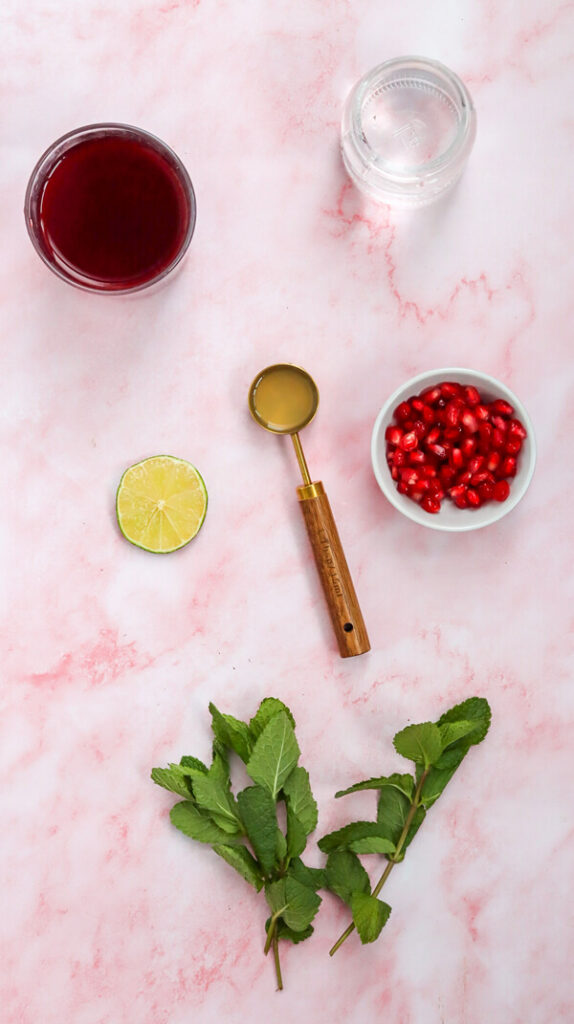 Kitchen counter top with pomegranate juice, pomegranate seeds, soda water, lime juice, a lime slice, and mint leaves