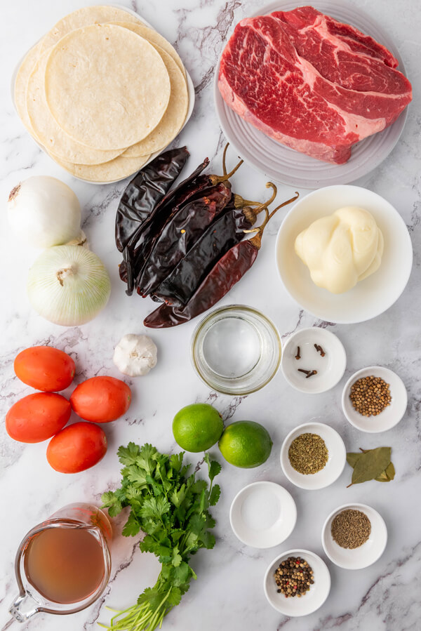 Counter top with tomatoes, onions, cilantro, limes, garlic, bay leaves, chilies and multiple bowls filled with tortillas, chuck roast, cumin, coriander, oregano, peppercorns, whole cloves, beef broth, water, and Oaxaca cheese