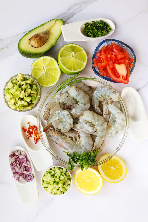 Kitcheb table with bowls of shrimps, cucumbers, onions, cilantro, jalapeño, and salt; next to some lemons, limes, and avocados