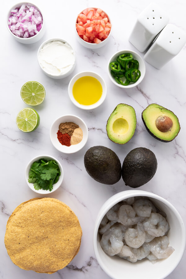 Multiple bowls filled with shrimps, tomatoes, onions, jalapeños, cilantro, olive oil, seasonings, and sour cream, next to limes, avocados, and tostadas