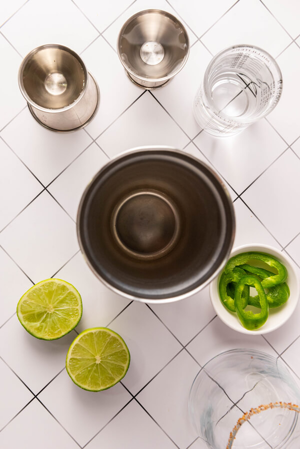 Cocktail shaker next to tequila, triple sec, agave syrup, limes, and jalapeno slices