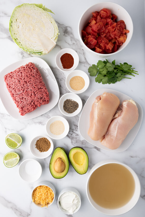 Counter top with cabbages, limes, avocados, cilantro, all next to some bowls with chicken stock, tomatoes, ground beef, chicken breasts, sour cream, shredded cheese, cumin, paprika, onion powder, garlic powder. black pepper, and salt