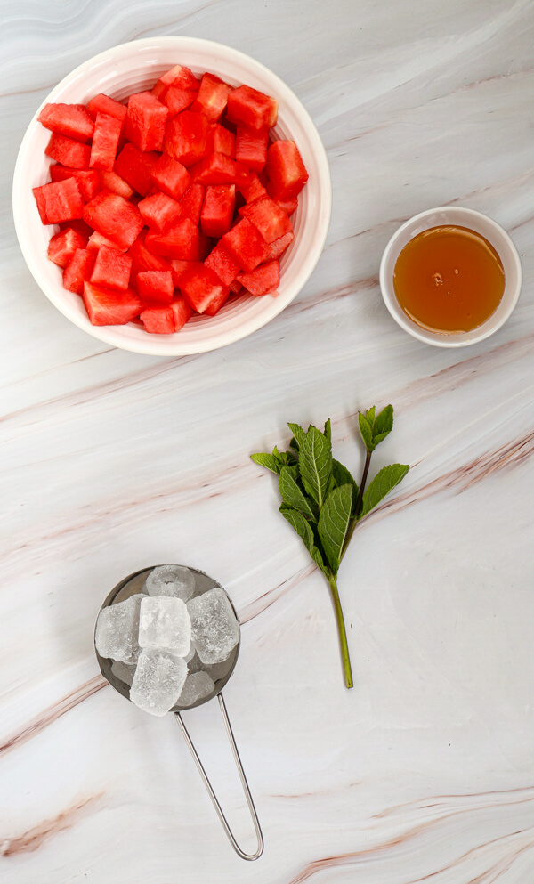 Kitchen counter top with a bowl of watermelon slices, a cup of honey, a cup of ice cubes, and some mint leaves
