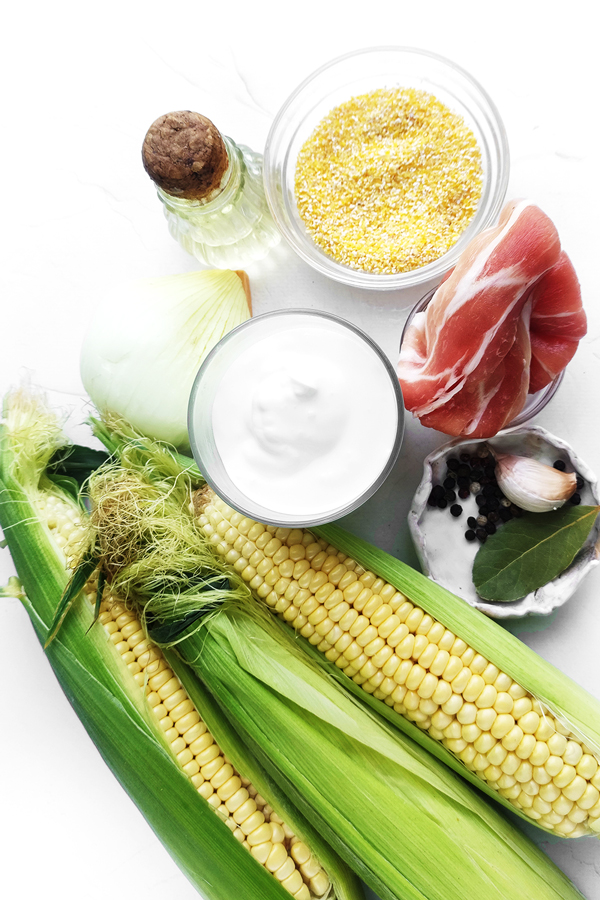 Table with corn cobs with husks, a bottle of olive oil, cups with cream, corn grits, bacon, peppercorns, bay leaves, garlic, and an onion 