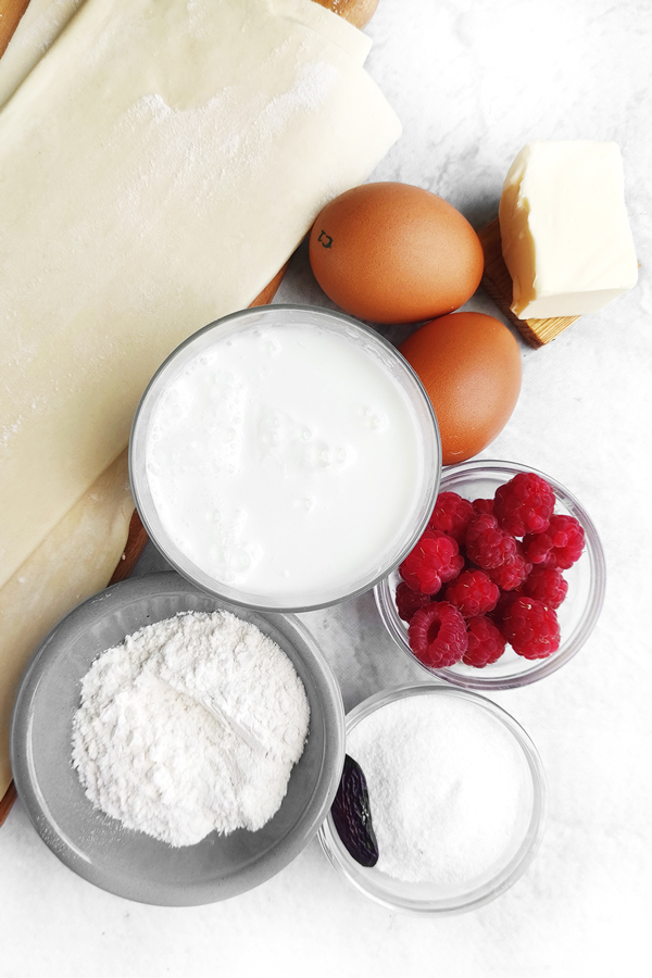 White table with puff pastry, eggs, cream, sugar, flour, tonka bean, and raspberries and butter