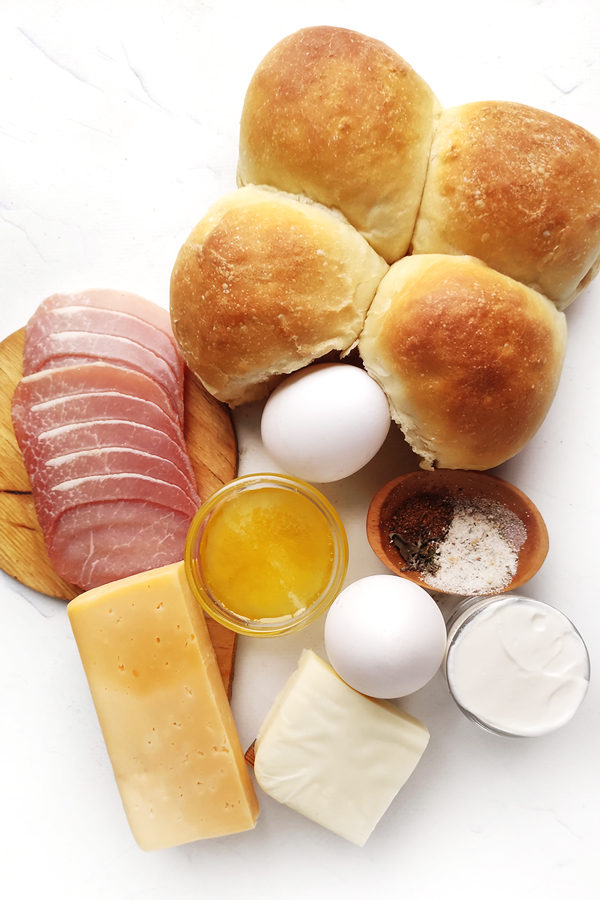 White table with 4 buns, ham, cheese, eggs, a cup of honey, a cup of cream, and seasoning