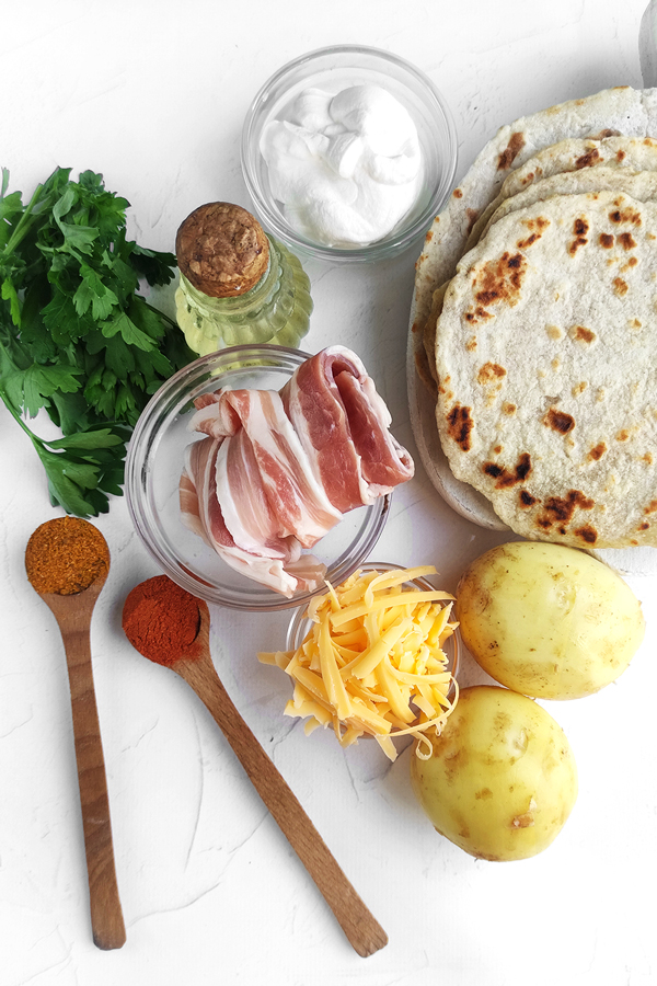 Kitchen counter top with potatoes, bacon, olive oil, tortillas, grated cheese, hers, a cup of sour cream, and two spoons with paprika and taco seasoning