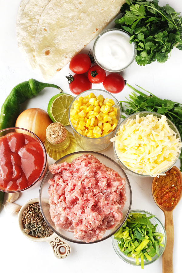 Kitchen table with tortillas, sour cream, tomatoes, cilantro, grated cheese, ground beef, corn, chilli pepper, onion, leeks, garlic, limes, olive oil, taco seasoning, and tomato sauce 
