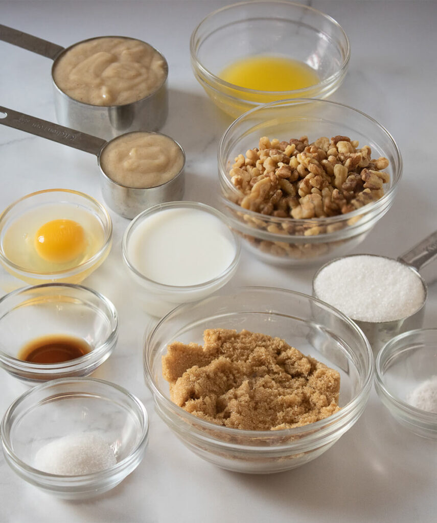 White table with bowls of mashed bananas, melted butter, milk, an egg, vanilla extract, brown sugar, granulated sugar, flour, salt, baking soda, and walnuts