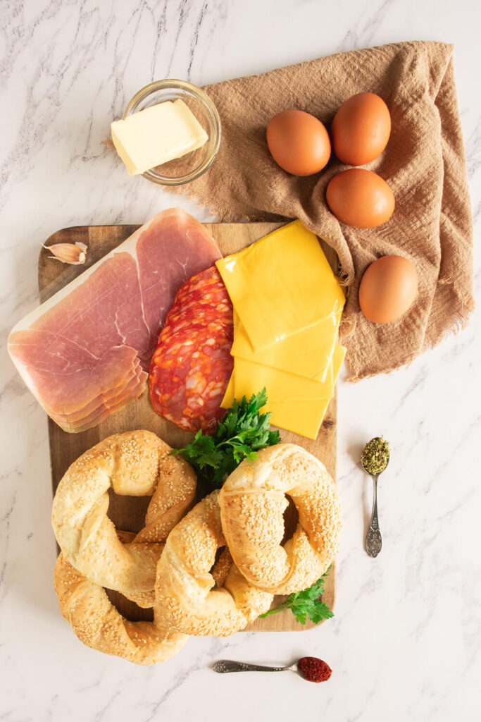Kitchen table with bagels, eggs, butter, cheese, salami, prosciutto, a garlic clove, dried oregano, paprika, and fresh parsley