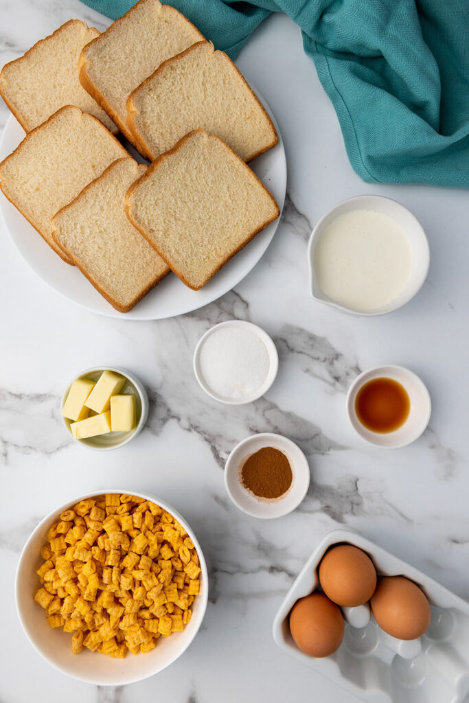 Kitchen counter top with a bowl of Cap'n Crunch cereal, slices of bread, eggs, butter, heavy cream, sugar, vanilla extract, and cinnamon