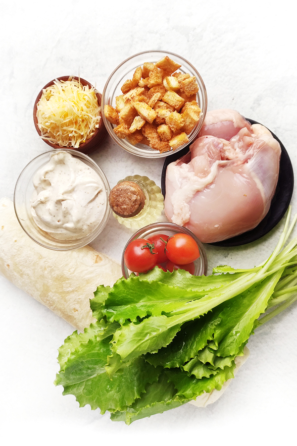 White table with tortillas, lettuce, tomatoes, chicken thighs, olive oil, Caesar dressing, croutons, and cheese