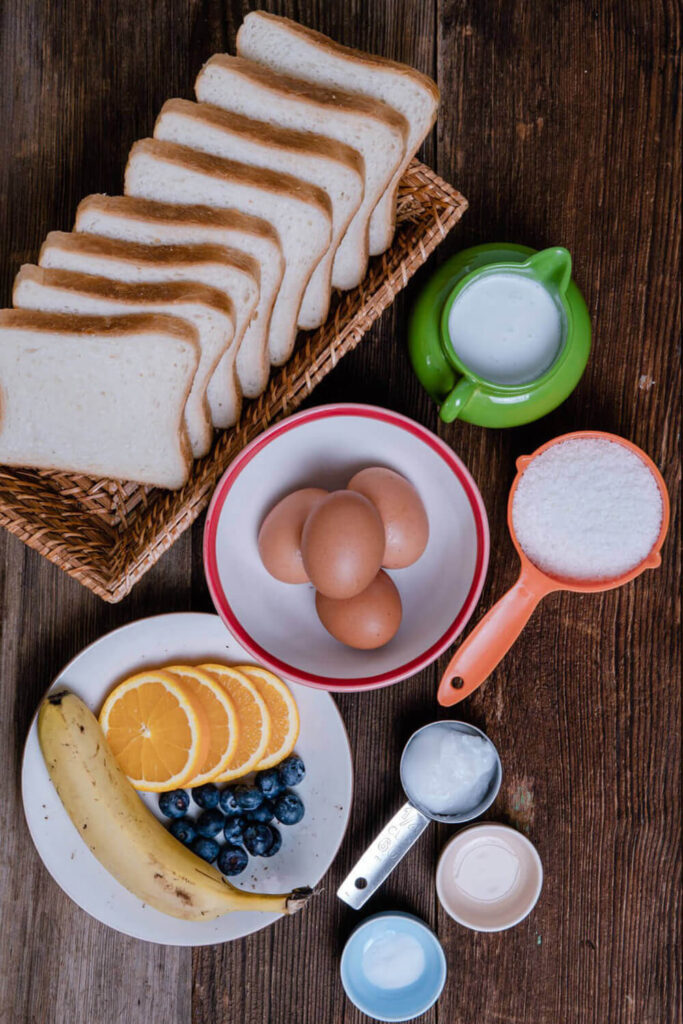 Wooden table with bowls of bread, eggs, coconut milk, shredded coconut, vanilla extract, coconut oil, salt, and fruits