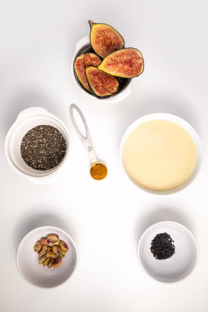 White table with cups of figs, oat milk, chia seeds, black sesame seeds, pistachios, and cinnamon