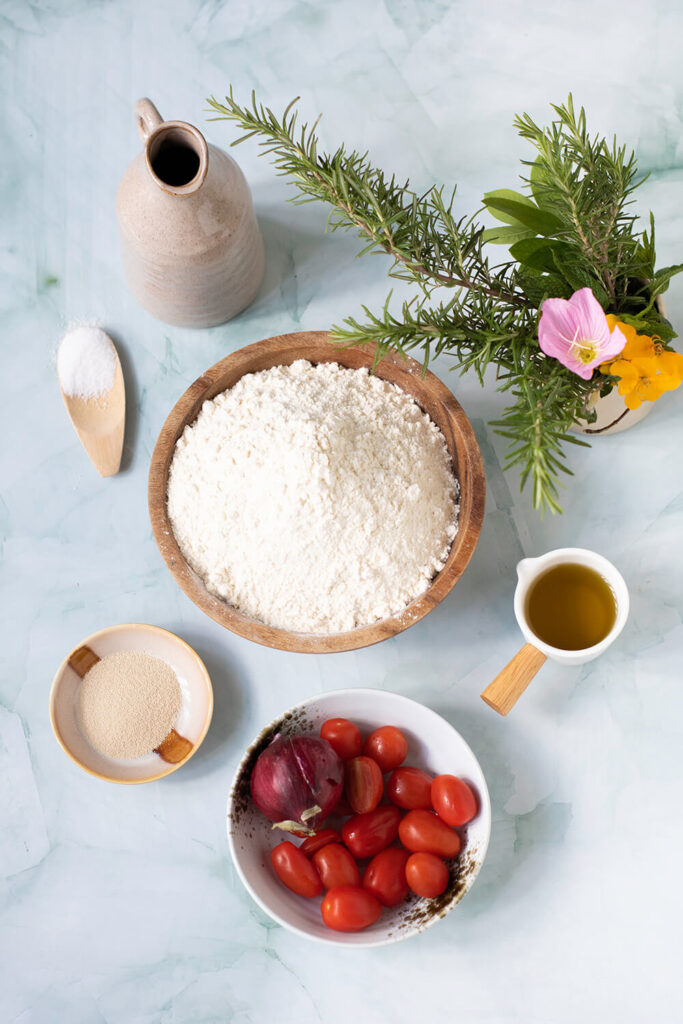 Blue table with a bowl of flour, a cup of yeast, a cup of olive oil, a spoon of sugar, a bowl with cherry tomatoes and an onion, and rosemary 