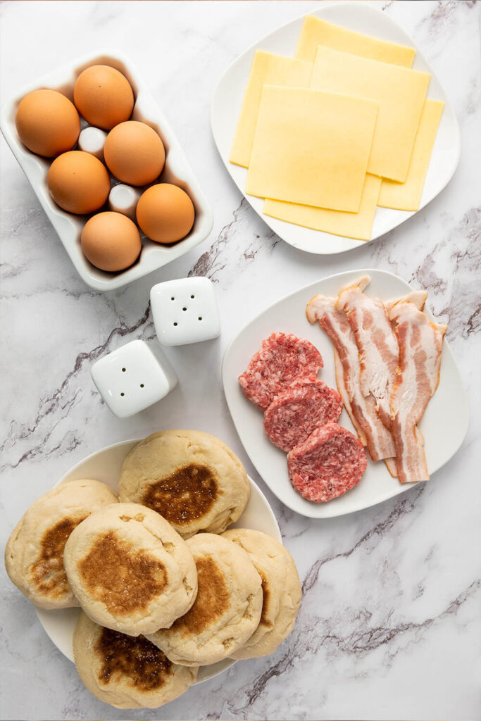 Kitchen counter top with eggs, cheese, bacon, sausages, English muffins, salt, and pepper