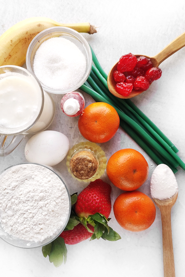 White table with cups of flour, buttermilk, sugar, baking powder, olive oil, an egg, food coloring, a banana, strawberries, 3 mandarins, cherries, and green licorice candies