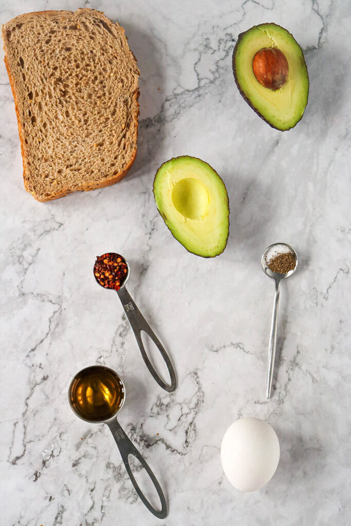 Marble table with bread slices, an avocado, an egg, and spoons of oil, chili, salt, and pepper