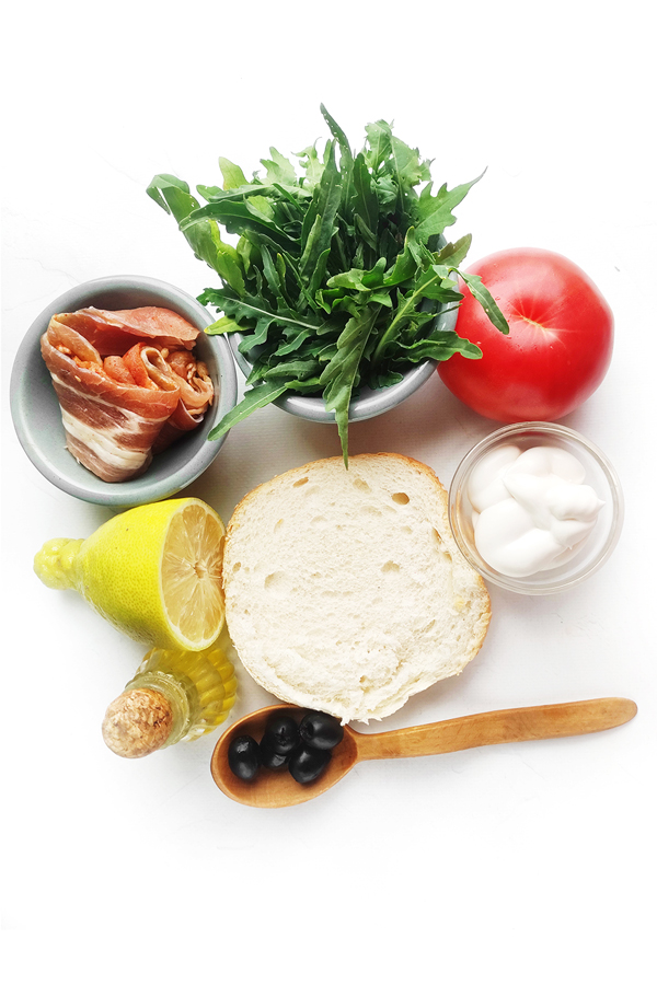 White table with half of a brioche, a tomato, a lime, arugula, bacon, mayonnaise, olives, and olive oil
