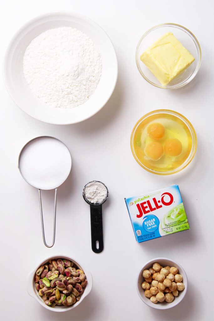 White table with bowls of flour, butter, sugar, baking powder, eggs, pistachos, hazelnuts, milk, and a box of pistachio pudding 