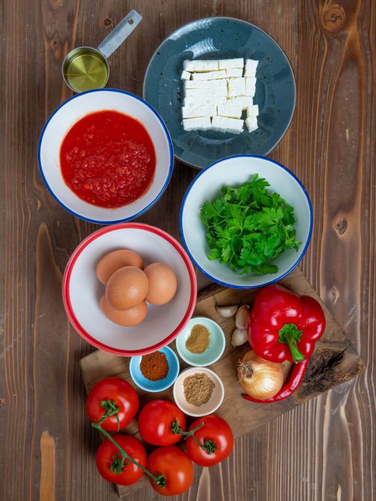 Wooden table with bowls of tomato passata, feta cheese, eggs, parsley, and oil, next to a cutting board holding tomatoes, an onion, a pepper, a chili, garlic cloves, and tiny cups of paprika, cumin, and coriander