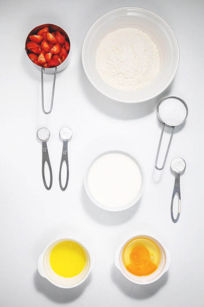 White table with bowls of flour, milk, strawberries, sugar, melted butter, eggs, and spoons of salt, baking powder, and baking soda