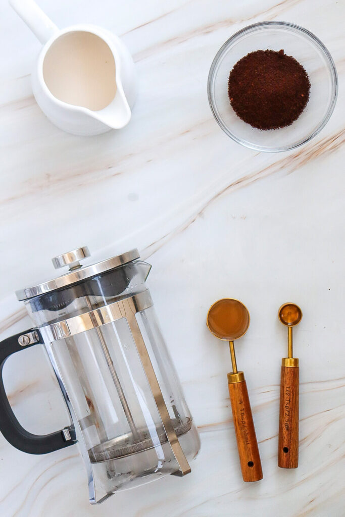 White marble countertop with a french press, a cup of ground coffee, a cup of milk, a spoon of caramel sauce, and half a  spoon of vanilla extract