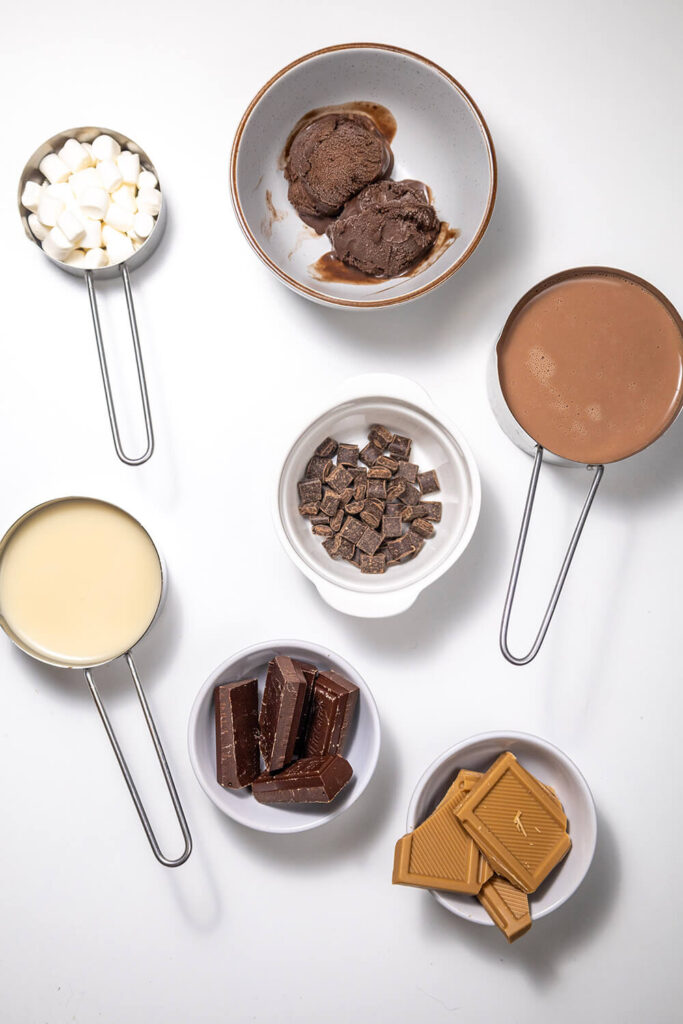 White table with bowls filled with chocolate ice cream, oat milk, chocolate milk, chocolate chips, chocolate bars, salted caramel chocolate bars, and marshmallows