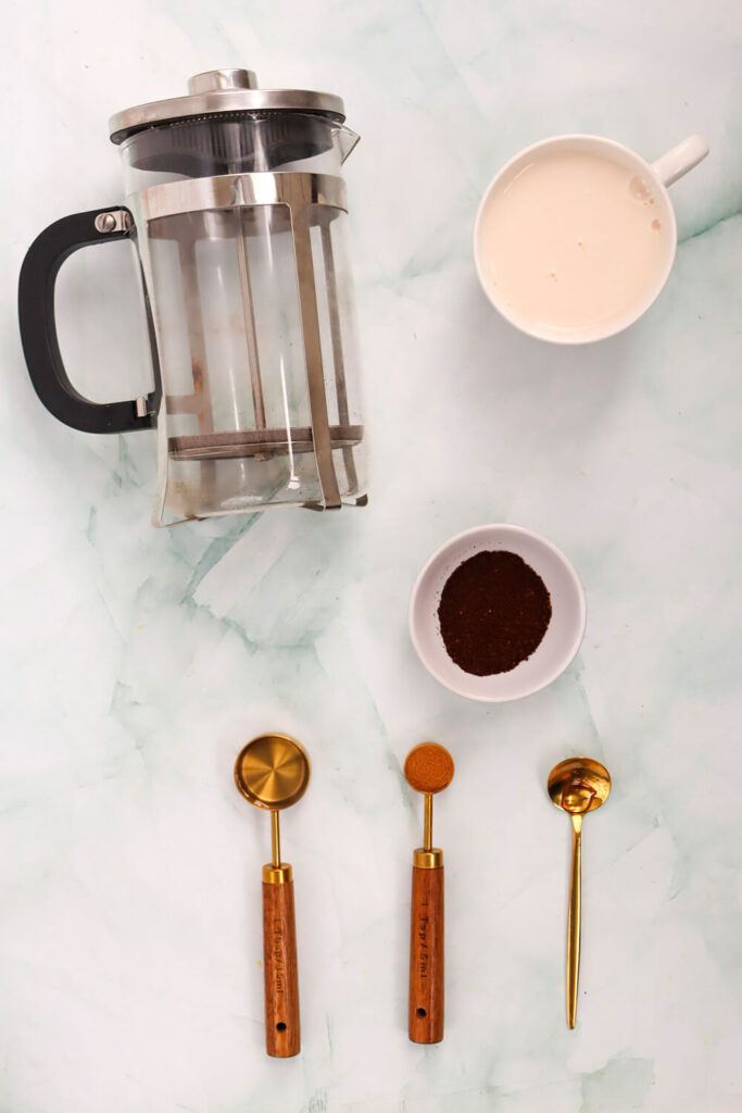 Blue marble countertop with a espresso machine, a cup of coffee, a cup of milk, and spoons of carob syrup, cinnamon, and vanilla extract