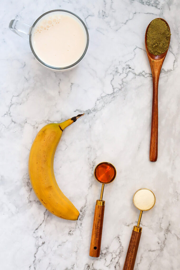 Marble countertop with a glass of almond milk, a spoon of green tea powder,a spoon of honey, a spoon of protein powder, and a banana 