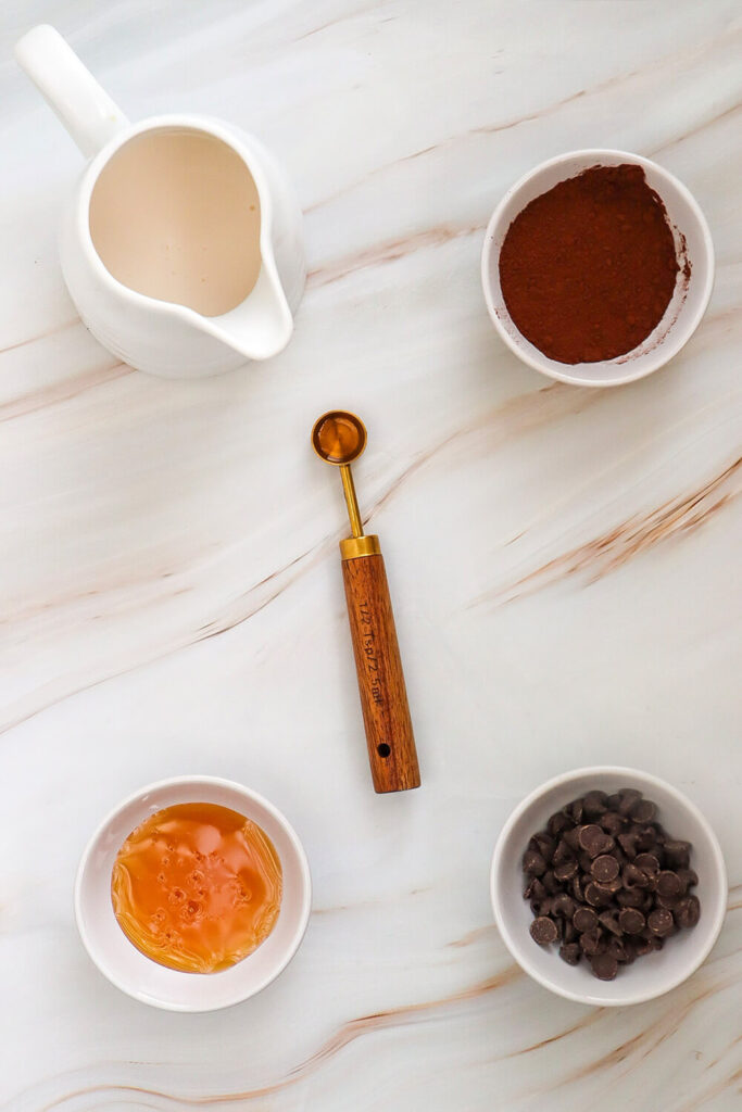 Marble countertop with bowls of almond milk, cocoa powder, maple syrup, chocolate chips, and vanilla extract