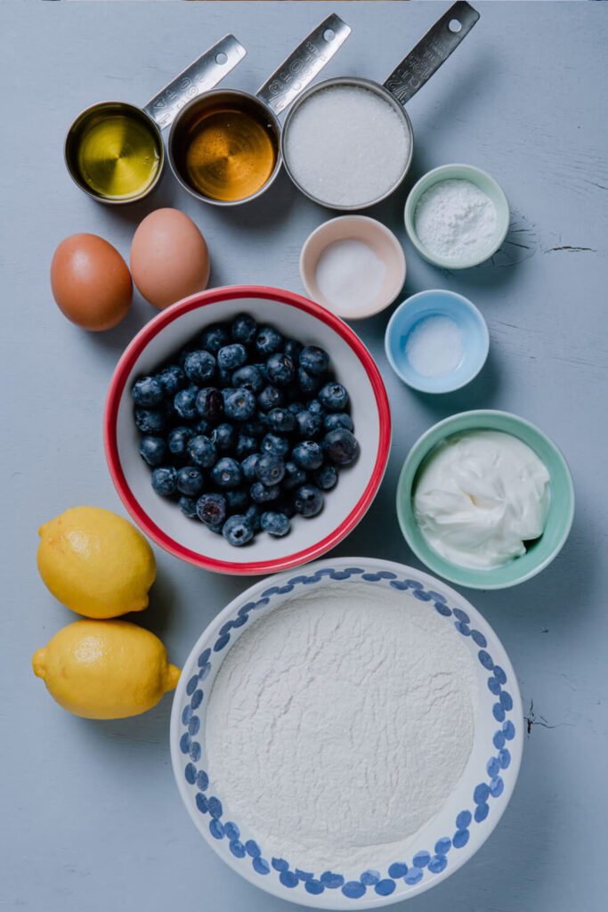 Blue table with bowl of blueberries, flour, baking powder, baking soda, salt, sugar, yogurt, oil, and honey, next to 2 eggs and 2 lemons