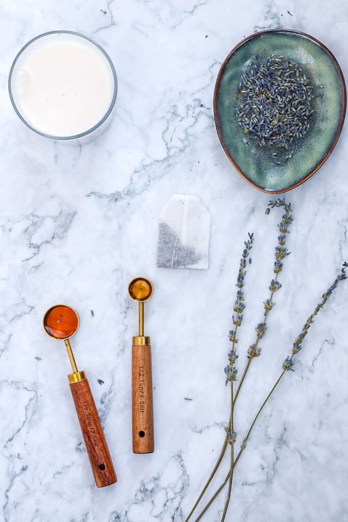 Marble countertop with bowls of milk and dried lavender, next to spoons of vanilla extract and honey, and a bag of Earl grey tea 