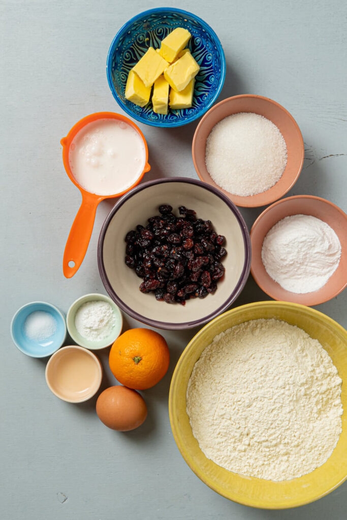 Concrete counter top with bowls of flour, sugar, cranberries, baking powder, salt, heavy cream, sour cream, vanilla extract, butter, an egg, and an orange