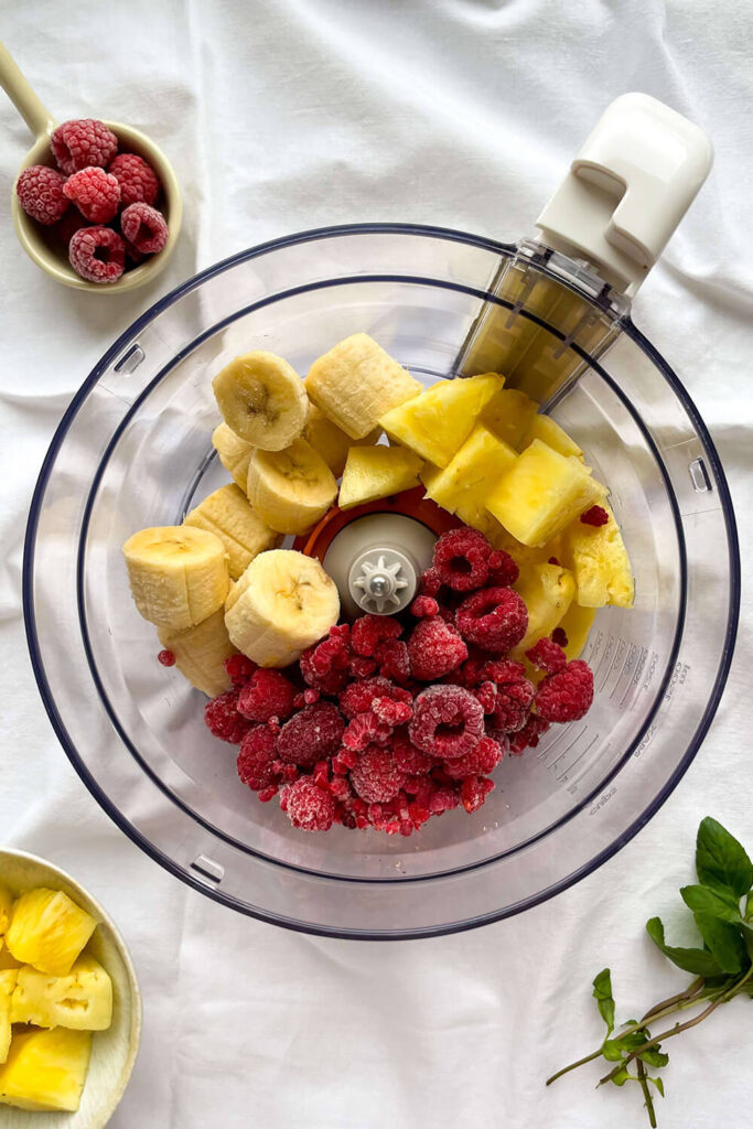 Adding the frozen bananas, raspberries, and pineapple to a food processor 