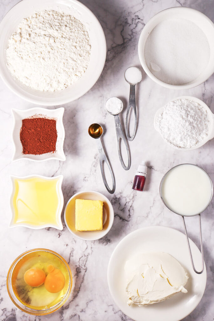 Marble counter top with bowls of flour, sugar, cocoa powder, baking soda, salt, oil, buttermilk, eggs, vanilla extract, red food coloring, cream cheese, butter, and powdered sugar