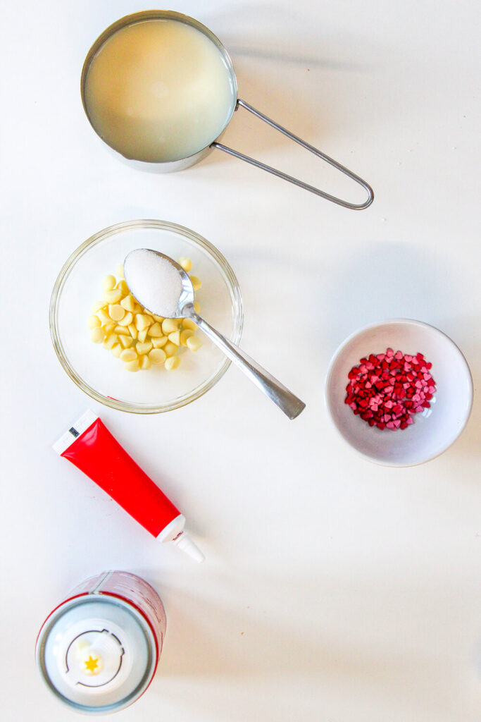 White kitchen table with bowls with milk, sugar, white chocolate chips, red food coloring, whipped cream, and red sprinkles