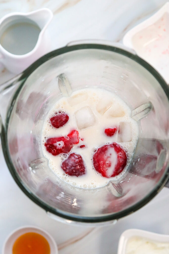 Blending almond milk, frozen strawberries, Greek yogurt, vanilla extract, cream cheese, and ice cubes