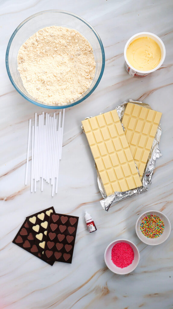 Marble table with bowls of cake mix, frosting and sprinkles, next to chocolate bars, red food coloring, and lollipop sticks
